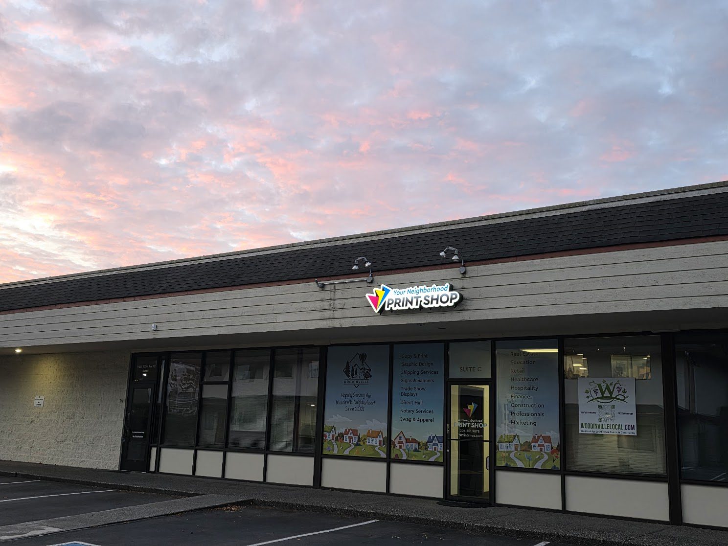 An outdoor daytime view of the storefront for Your Neighborhood Print Shop in Woodinville, Suite C. The windows display a list of services including Graphic Design, Shipping, and Direct Mail, alongside a 'Happily Serving the Woodinville Neighborhood' graphic.