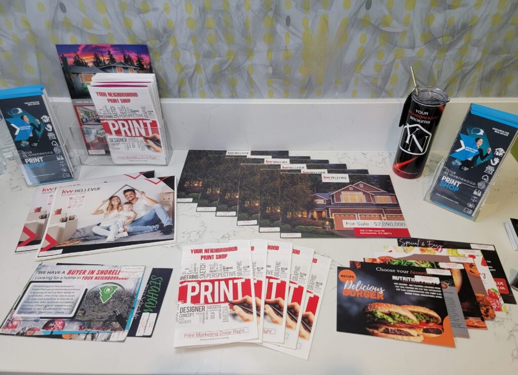 A large spread of custom Marketing Materials from Your Neighborhood Print Shop, including real estate 'For Sale' postcards, restaurant menu flyers, promotional booklets, a branded water bottle, and vertical banner stands.